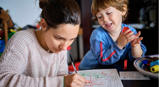 : A woman in a cable-knit sweater draws a red figure on paper while a young boy watches with a smile.